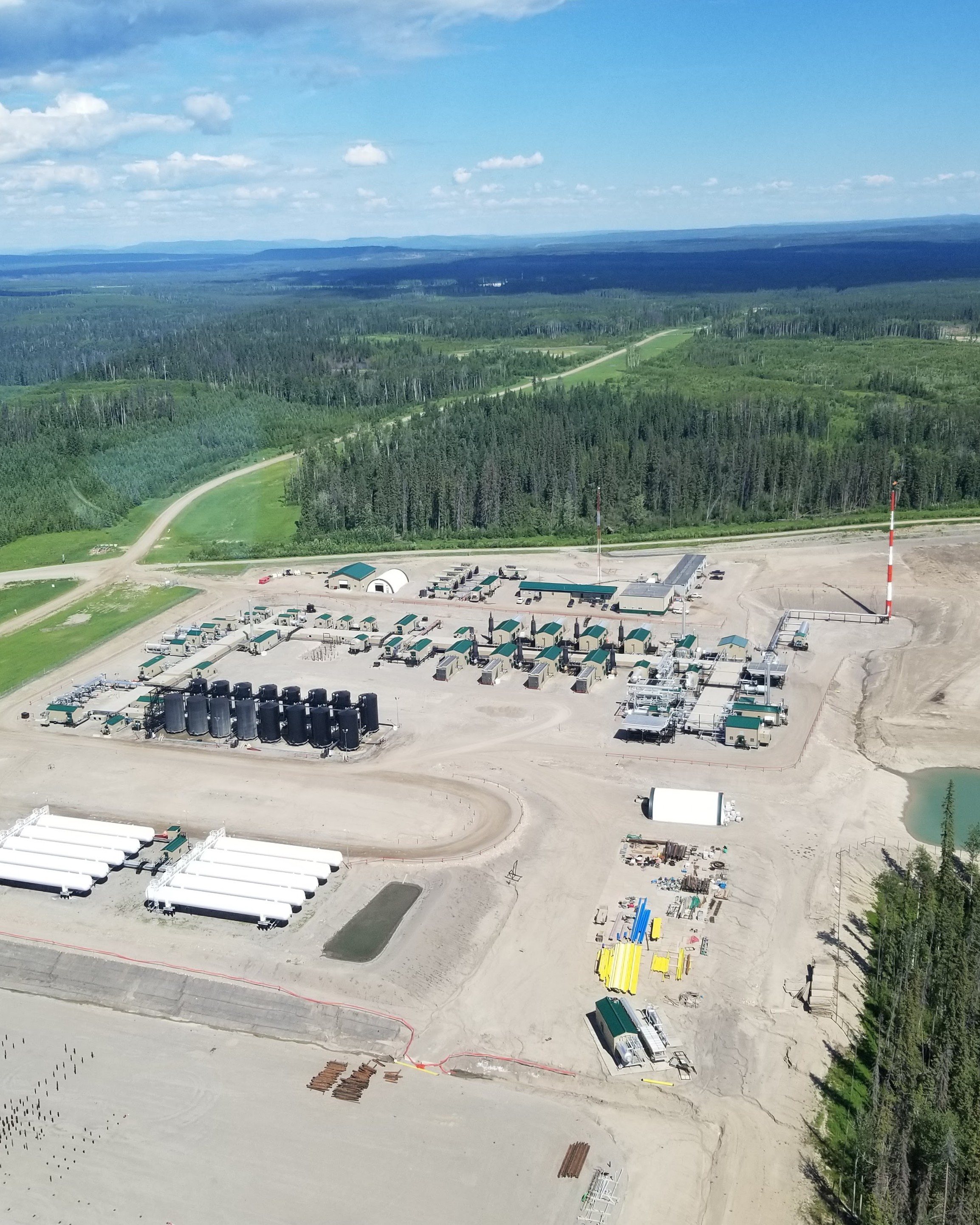 Aerial view of a remote gas processing facility with green-roofed buildings, storage tanks, and a flare stack surrounded by boreal forest