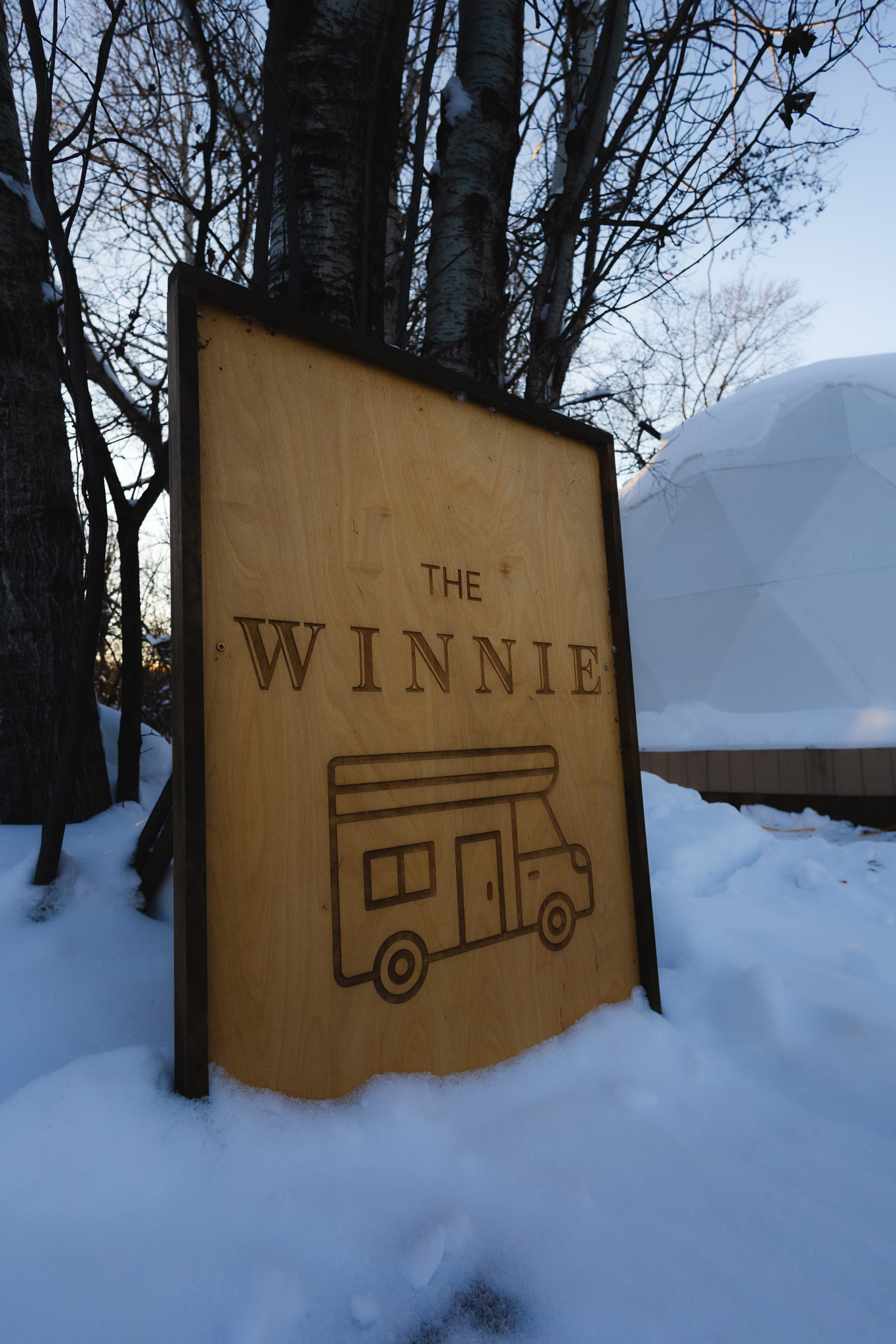 Engraved 'The Winnie' camper sign in the snow beside a geodesic dome