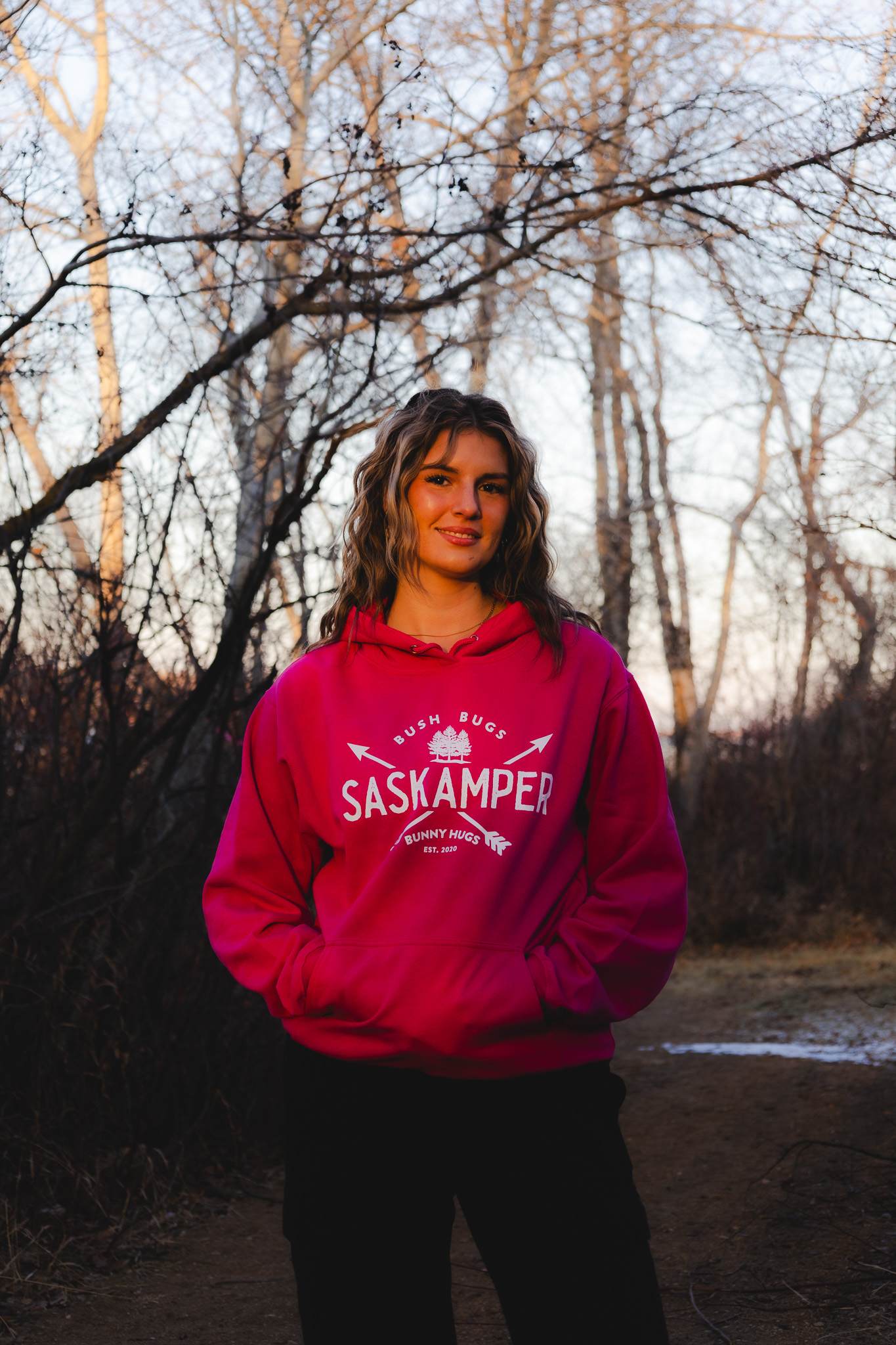 Woman in a pink Saskamper hoodie in golden-hour woods