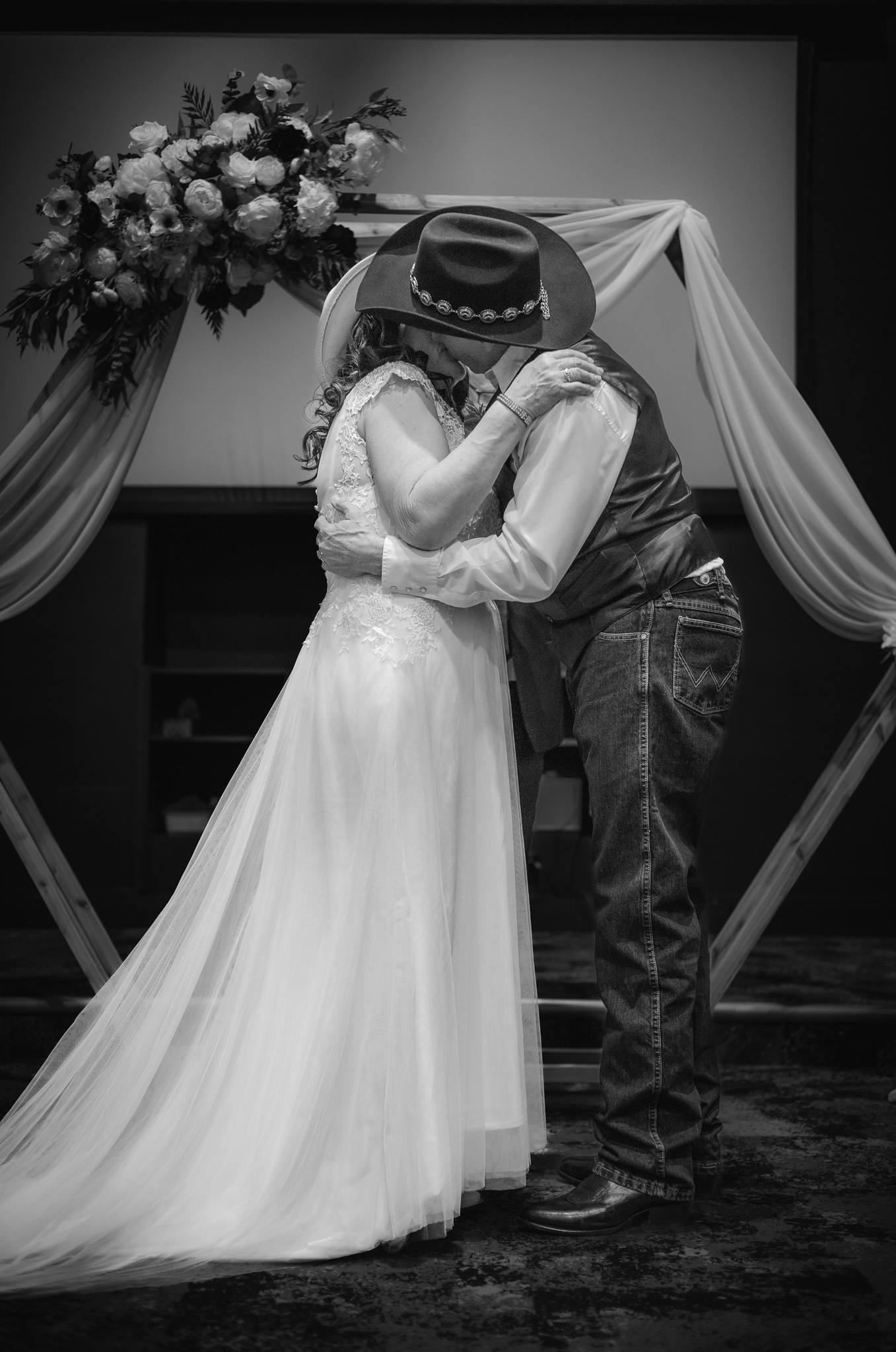 Black and white photo of a cowboy-themed couple's first kiss under a floral arch